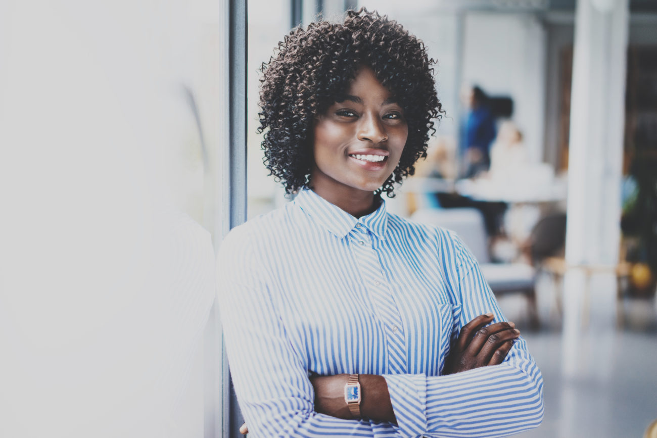 Portrait of happy beautiful african woman standing with arms folded and colleagues on background in modern loft.Businesswoman looking at the camera. Horizontal,blurred.